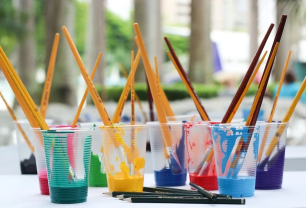 Various plastic cups with different colour paints in them, alongside other artistic equipment