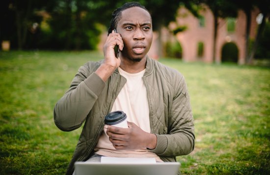 Stock image of a young black man speaking on the phone, while sitting outside with coffee in his hand.